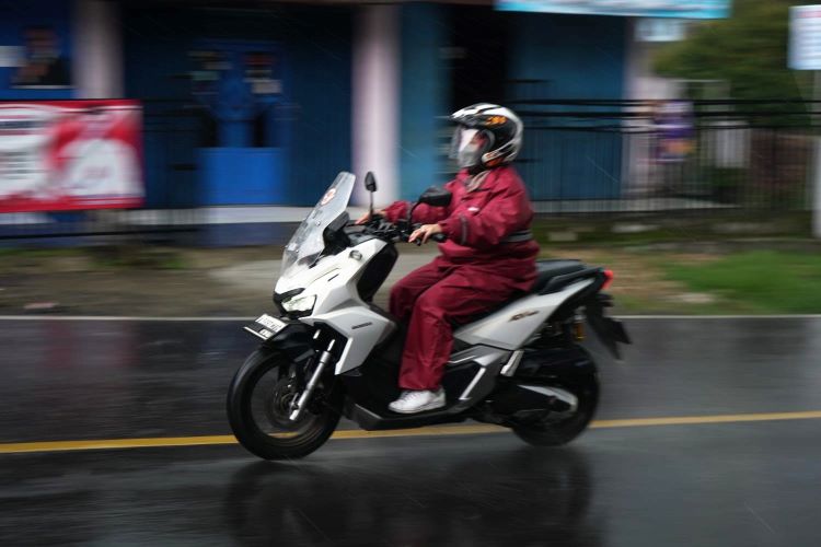 Seorang wanita yang mengendarai motor Honda ADV mengenakan jas hujan yang tepat saat hujan